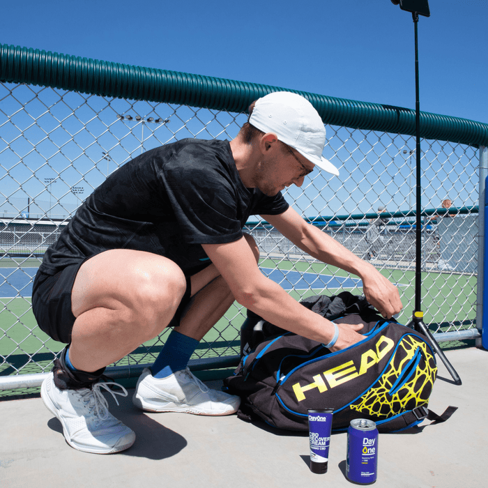 Man crouching on the ground outside pickleball court packing his pickleball bag with Day One recovery cream and CBD sparkling water on the side
