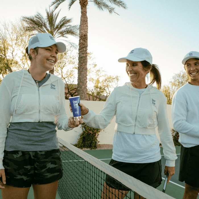 Three people wearing white Day One jackets on a pickleball court holding Day One recovery cream