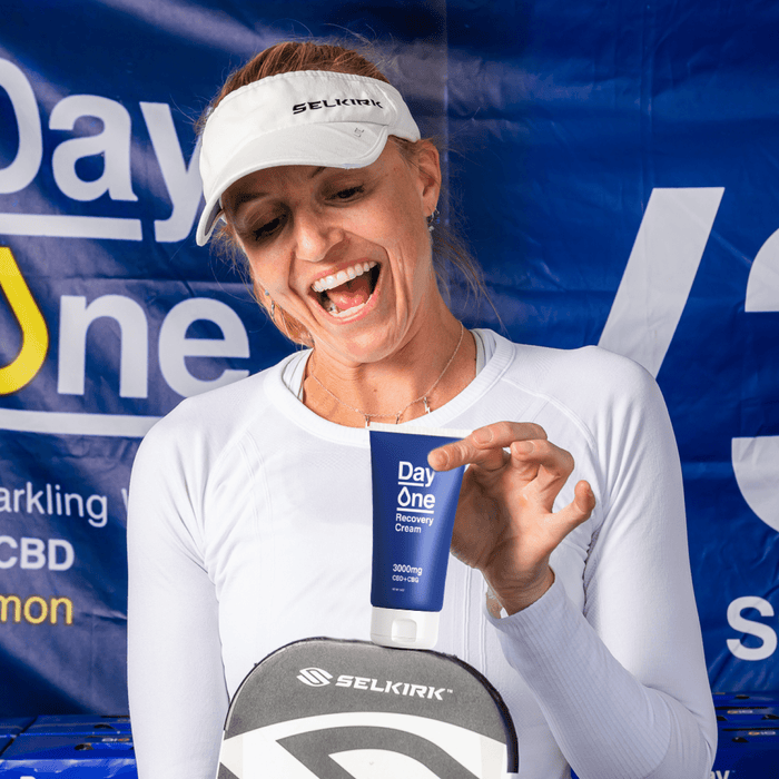 Woman in white sports clothes holds Day One recovery cream above Selkirk pickleball paddle 